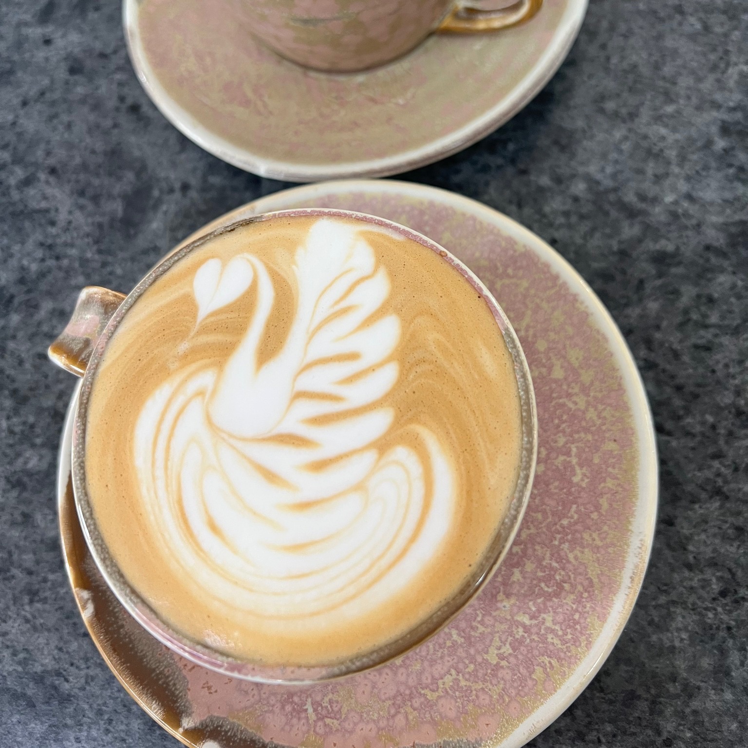 A flat white with a swan poured into it, in a glazed pink cup and saucer, shot from above on a dark stone table.
