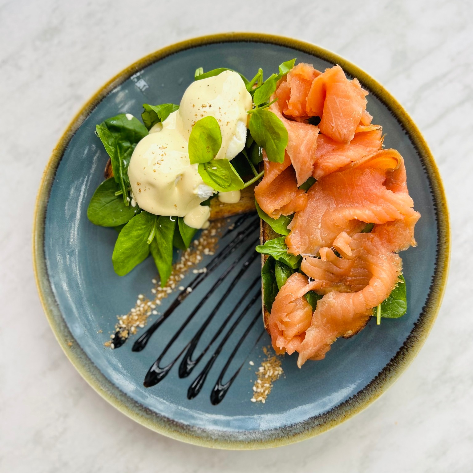 Smoked salmon and a poached egg under hollandaise on a bed of greens, finished with balsamic and sesame, on a blue-rimmed plate.