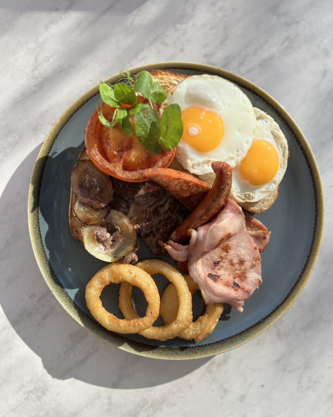 A big breakfast on a blue-rimmed plate: two fried eggs, bacon, mushrooms, grilled tomato and onion rings on toast, shot from above on marble.
