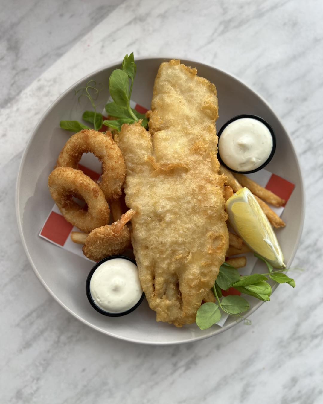 Battered fish, calamari rings, chips, a lemon wedge and two pots of tartare on a pale plate, shot from above on marble.