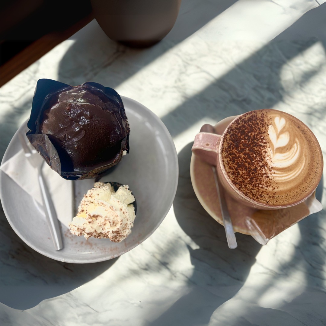 A chocolate-topped muffin and a spoonful of cream on one plate and a cappuccino on another, on a marble table with morning light falling across them in patches.